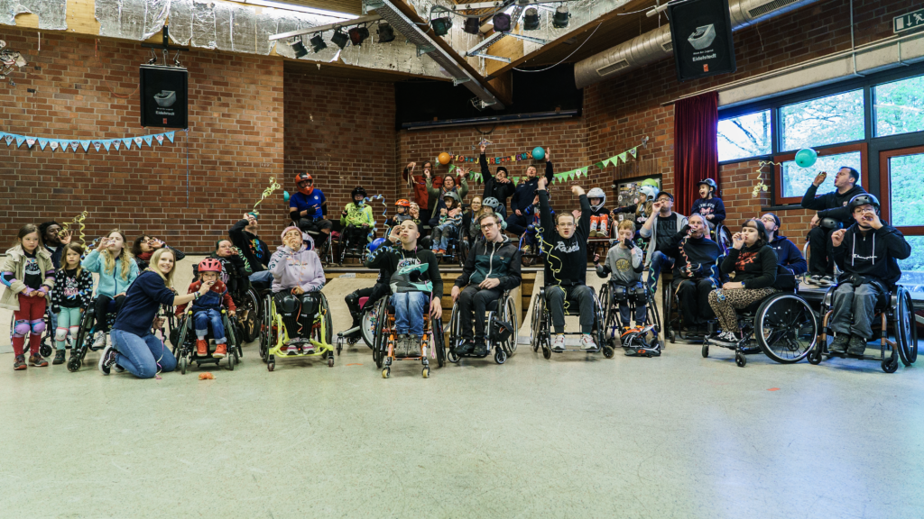 Auf einem Gruppenfoto in einer Skatehalle stehen viele Menschen, vorrangig Rollstuhlfahrer und Rollstuhlfahrerinnen unterschiedlichen Alters vor und auf Rampen. Sie jubeln und pusten Luftschlangen in die Luft.