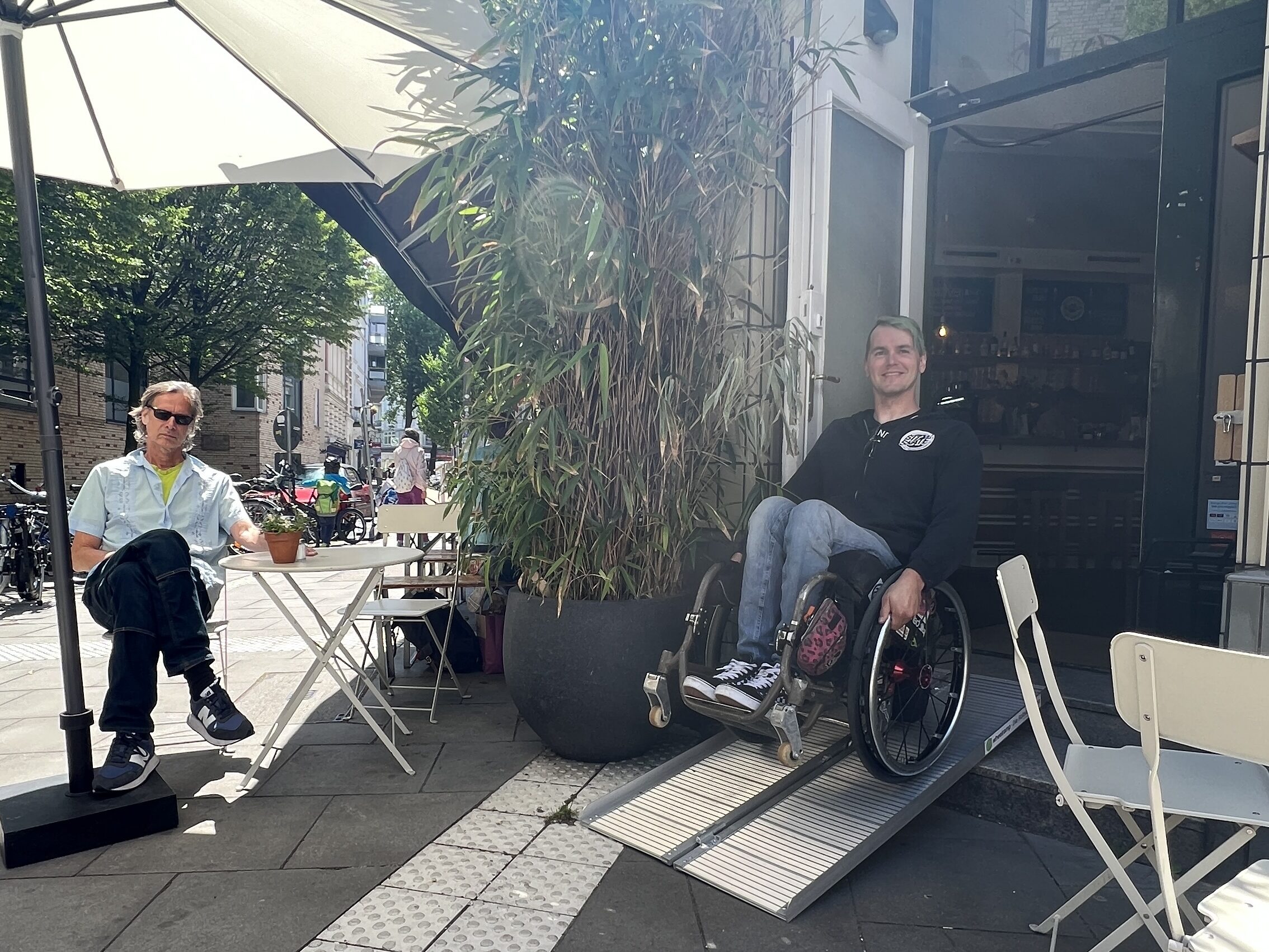 Ein Rollstuhlfahrer auf einer Rampe am Eingang eines Cafés. Daneben sitzt ein Mann an einem Tisch draußen vor dem Laden. 