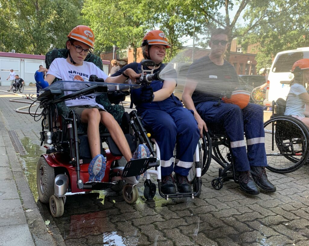 Drei Rollstuhlfahrer, davon zwei im Feuerwehranzug spritzen Wasser aus einem Feuerwerhschlauch.