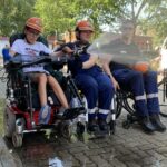Drei Rollstuhlfahrer, davon zwei im Feuerwehranzug spritzen Wasser aus einem Feuerwerhschlauch.