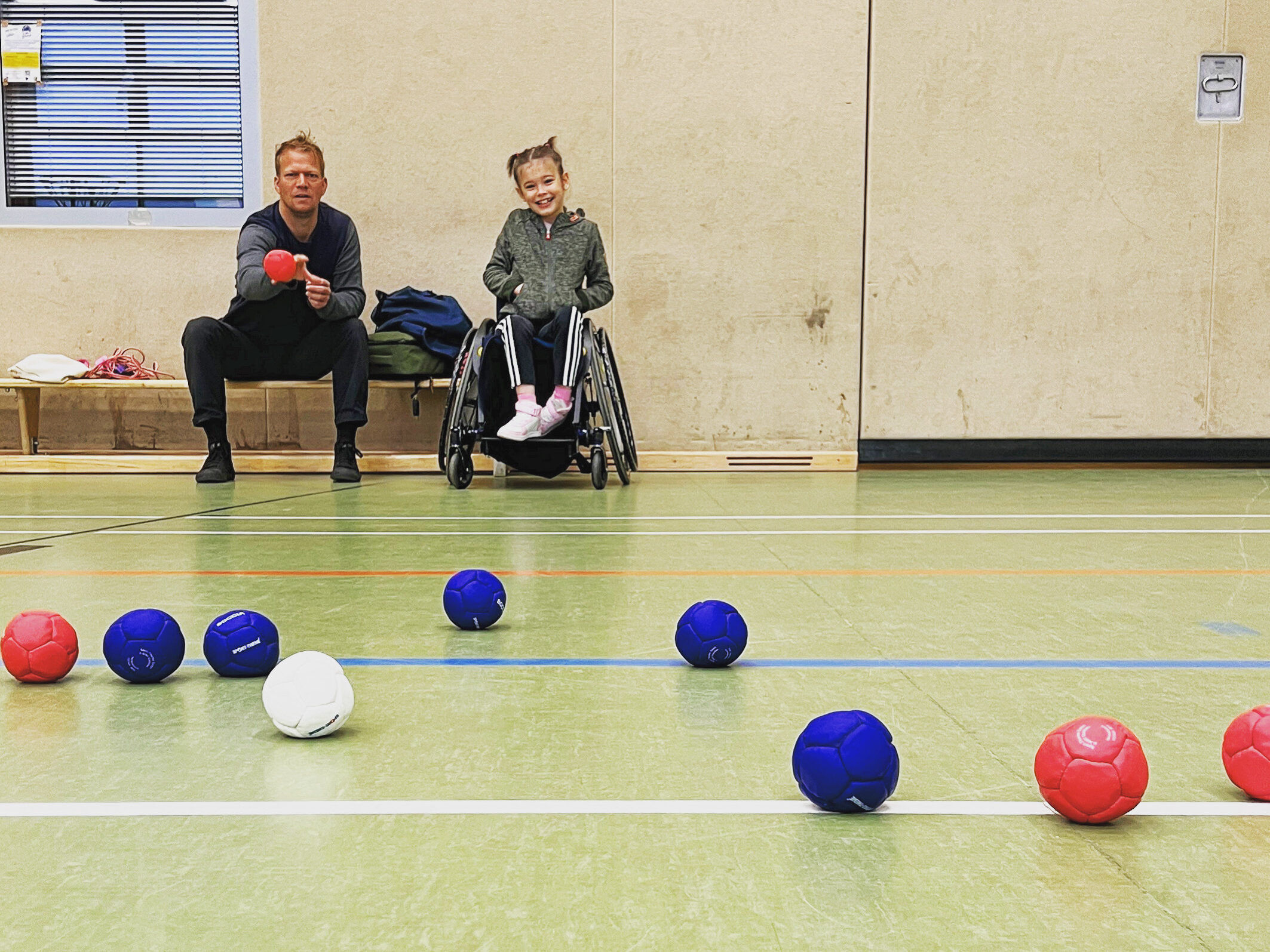  In einer Sporthalle spielen ein Mädchen im Rollstuhl und ein Mann, der auf einer Bank neben dran sitzt, eine Runde Hallen Boccia. Dabei werfen Sie blaue und rote Bälle in Richtung eines weißen Balls.