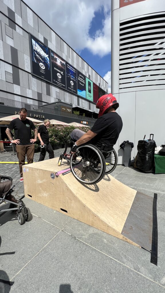 Ein Mann im Rollstuhl macht einen Trick auf einer Skaterampe aus Holz, die auf einen urbanen Platz gestellt wurde.