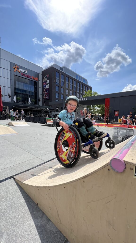 Ein Kind im Rollstuhl auf einer Skaterampe aus Holz, die auf einen urbanen Platz gestellt wurde.