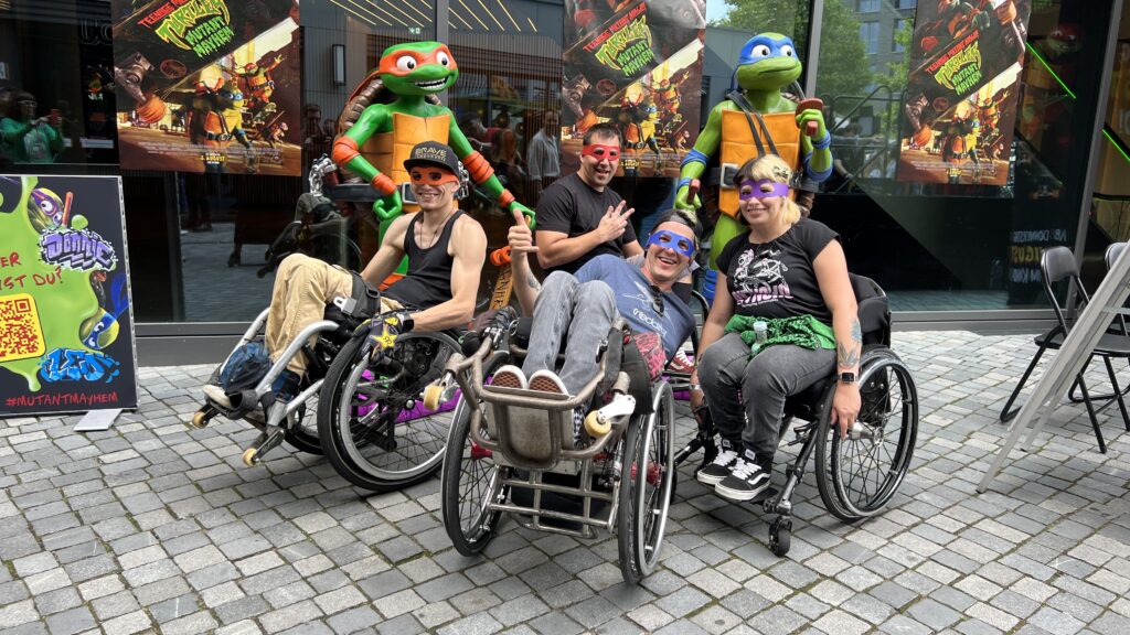 Drei Männer im Rollstuhl und eine Frau im Rollstuhl stehen vor zwei Figuren der Teenage Mutant Ninja Turtles. Sie haben die bunten Augenbinden der Turtles auf. 