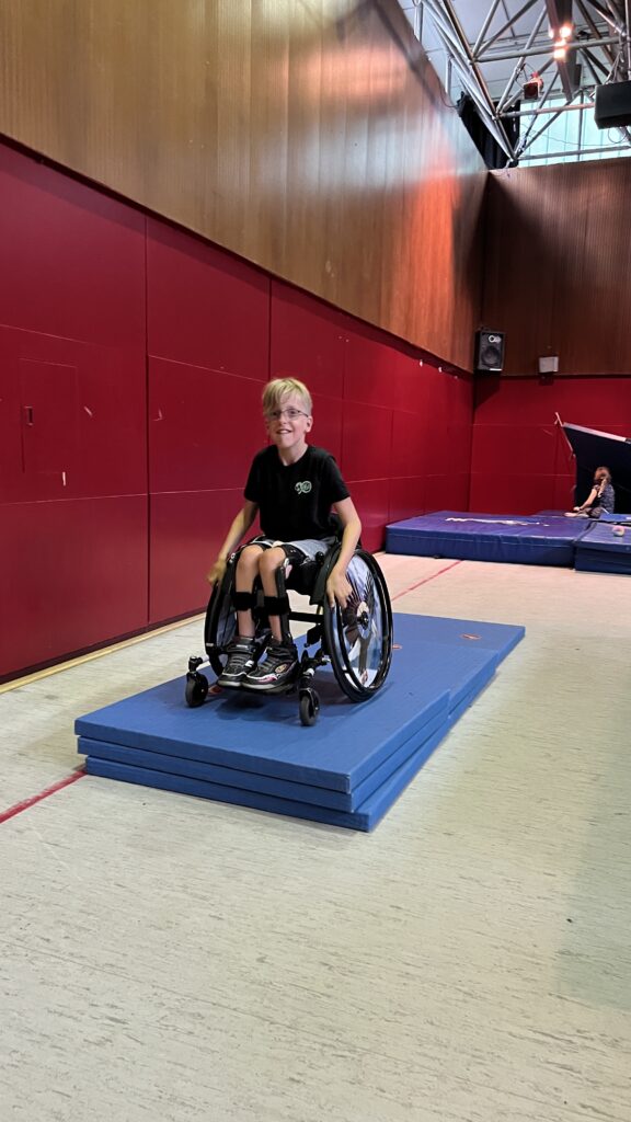 Ein Junge im Rollstuhl fährt in einer Sporthalle über aufgetürmte Matten.
