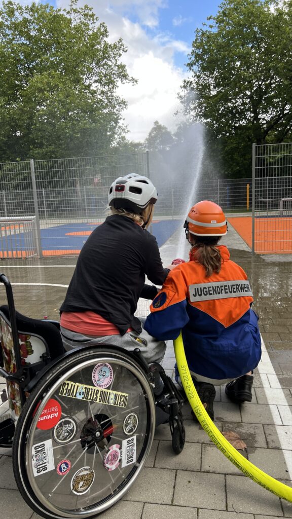 Eine Rollstuhlfahrerin und eine Frau in Feuerwehrkleidung auf der Jugendfeuerwehr steht, halten gemeinsam einen Schlauch aus dem Wasser gespritzt wird.