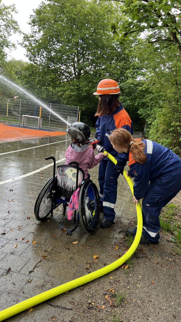 Ein Kind im Rollstuhl hält einen Wasserschlauch zusammen mit zwei Jugendlichen in Feuerwehr Jacken. 
