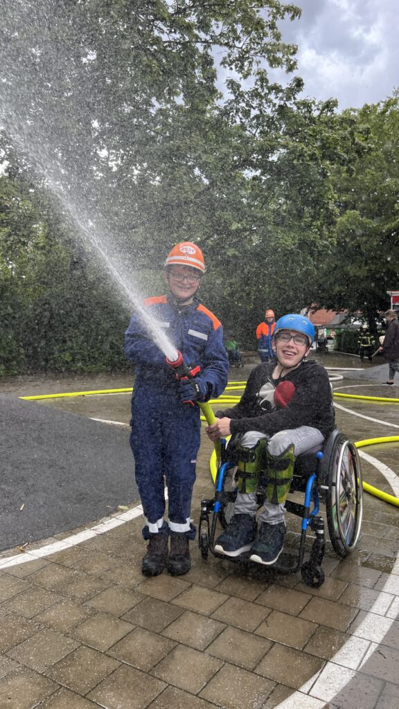 Ein jugendlicher Rollstuhlfahrer und ein Jugendlicher in Feuerwehruniform halten einen Wasserschlauch aus dem ein Wasserstrahl kommt. 