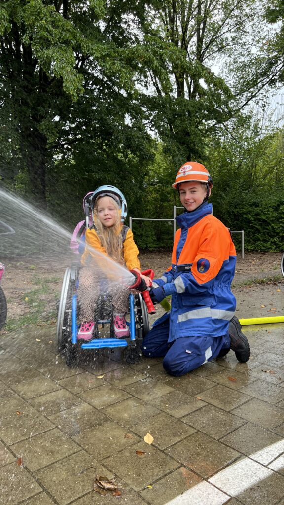 Ein Jugendlicher in Feuerwehrjacke und ein Mädchen im Rollstuhl halten einen Feuerwehrschlauch.