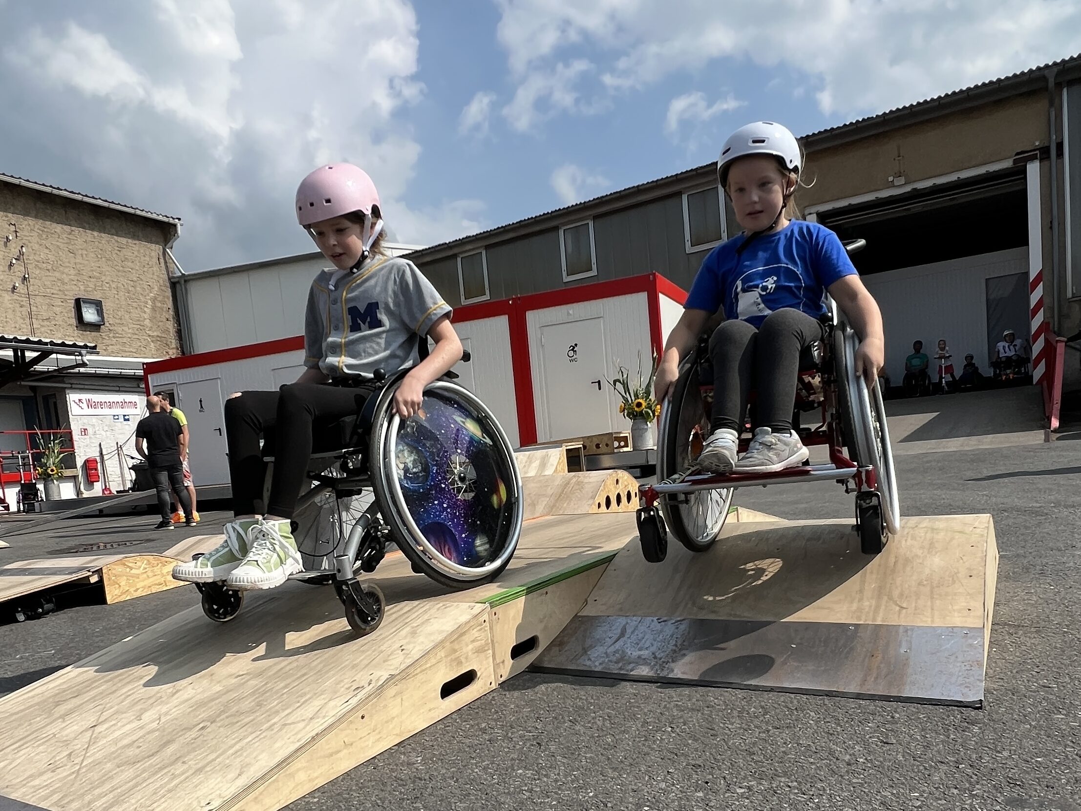 Zwei Mädchen im Rollstuhl fahren nebeneinander über zwei Skateelemente aus Holz, die auf einem geteerten Platz gestellt wurden.