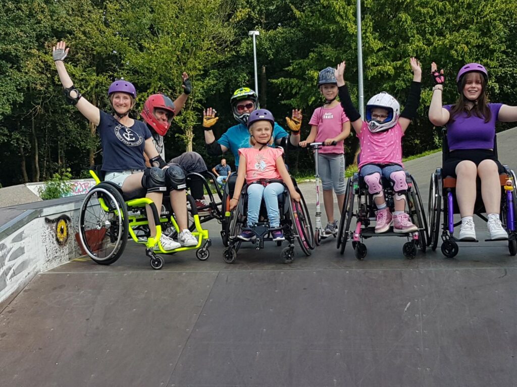 Eine Gruppe von Rollstuhlfahrerinnen und ein Rollstuhlfahrer stehen im Skatepark auf einer Rampe für ein Gruppenfoto zusammen. Einige jubeln.