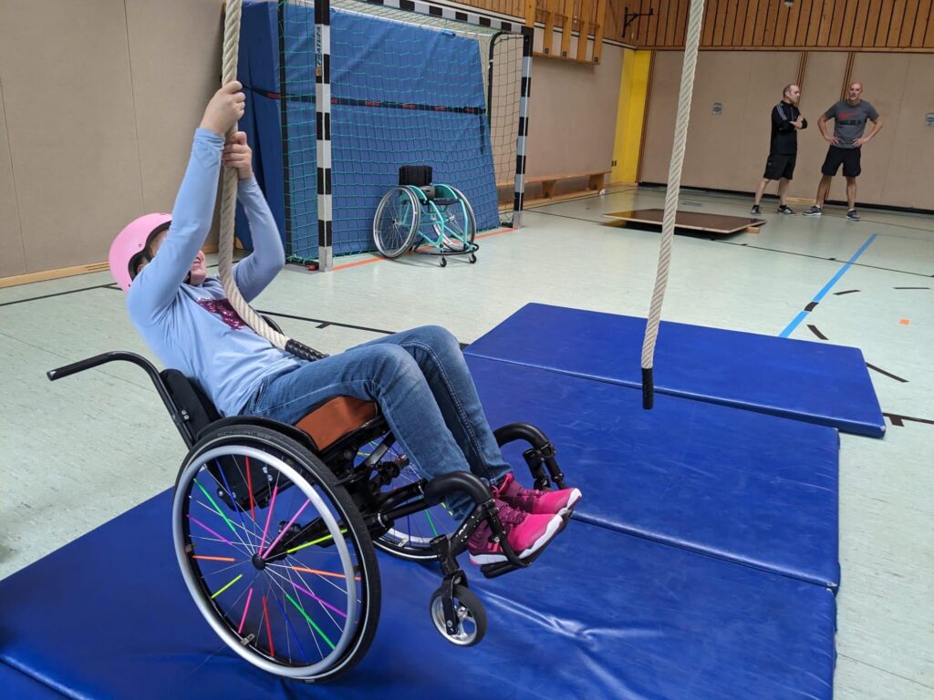 in Mädchen im Rollstuhl in einer Sporthalle zieht sich an einem Seil hoch. Der Rollstuhl ist dabei leicht nach hinten gekippt.