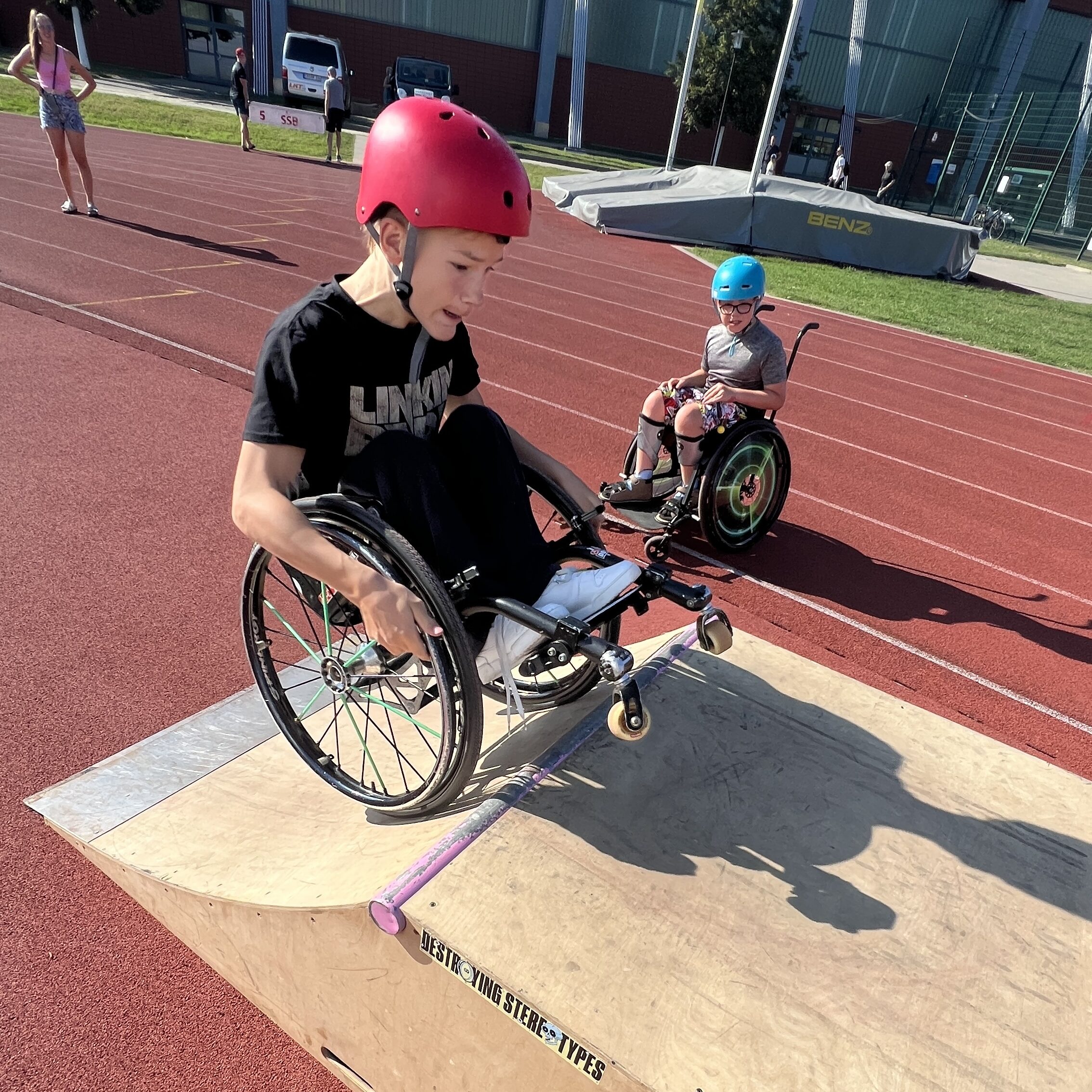 Ein Junge im Rollstuhl macht einen Trick auf einer mobilen Skaterampe, die auf einer Leichtathletikbahn steht. Daneben staunt ein weiterer Junge im Rollstuhl.