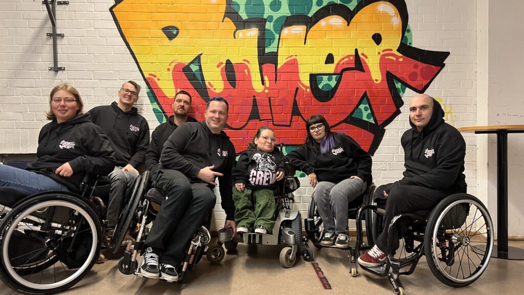 Rollstuhlfahrer und Rollstuhlfahrerinnen stehen zusammen, einige schräg angekippt, andere zurückgelehnt. In der Mitte steht eine Rollstuhlfahrerin im Elektrorollstuhl. Alle haben ein Crew Pullover an. An der Wand ein Graffiti mit dem Wort power.