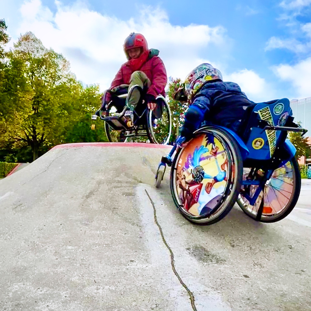 Zwei Rollstuhlfahrer im Skatepark an einem Vulkanförmigen Obstacle. Ein erwachsener Rollstuhlfahrer balanciert auf den Hinterrädern auf dem Vulkan, während ein Junge im Rollstuhl an der Seite des Vulkans hoch fährt.