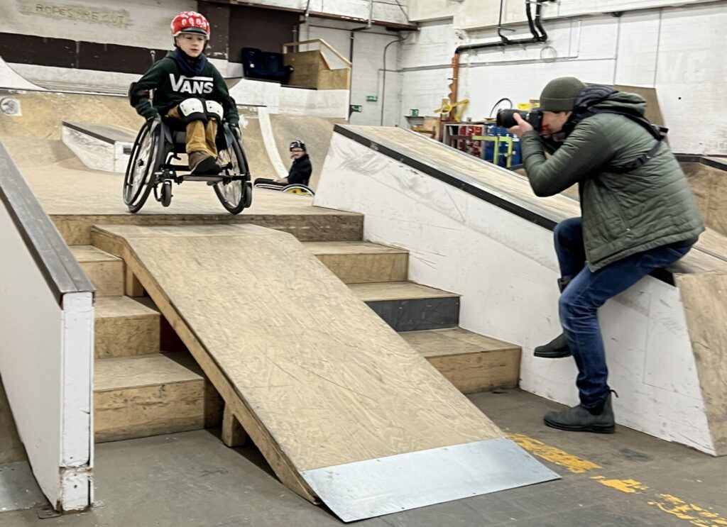 Bild von einem Kind im Rollstuhl in einer Skatehalle auf den Hinterrädern balancierend auf einer Rampe.