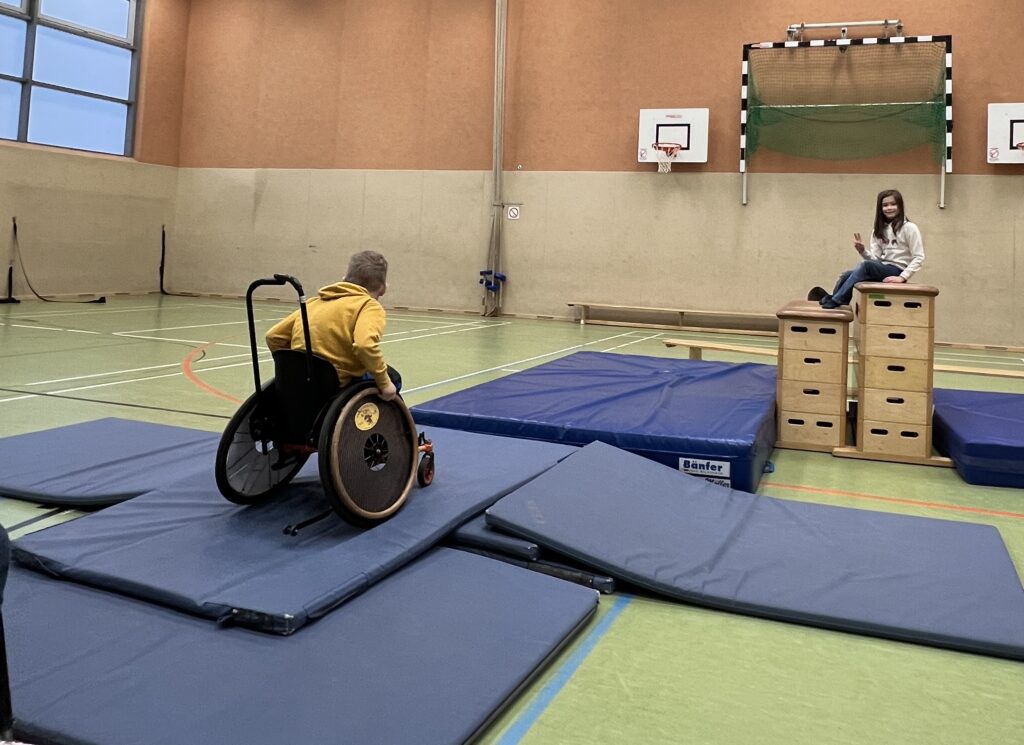 In einer Sporthalle fährt ein Junge im Rollstuhl über aufeinander gelegte Turnmatten. Im Hintergrund sitzt ein Mädchen auf Kästen. 