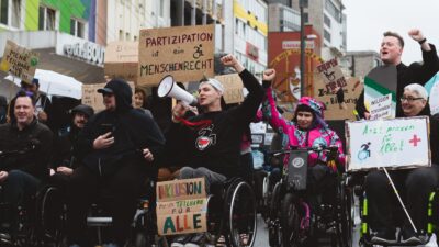 Eine Gruppe von Menschen, viele davon im Rollstuhl, nimmt an einer Demonstration teil. Sie halten Schilder und Transparente hoch. Im Vordergrund ist ein großes Transparent mit der Aufschrift "Inklusion für alle" zu sehen.