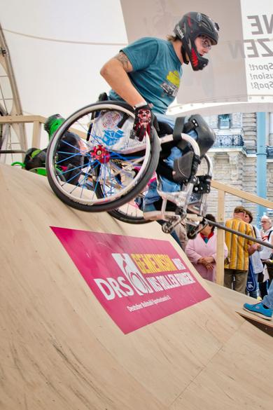 Ein Rollstuhlfahrer fährt in einer Messe halle von einer Skaterampe.