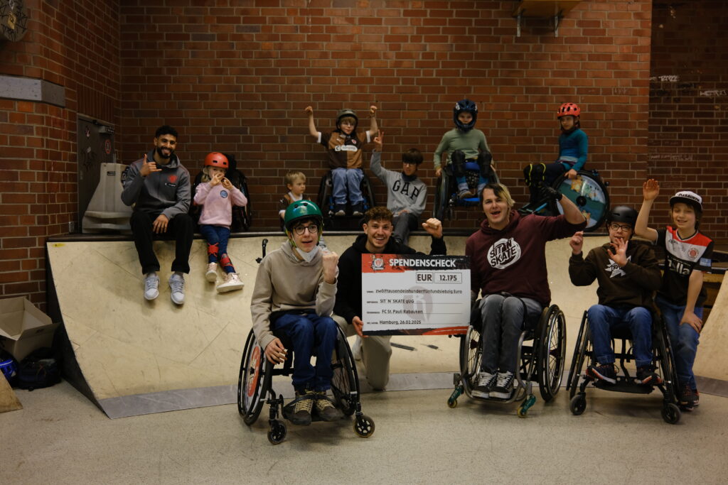 Eine große Gruppe von Kindern und Jugendlichen, viele davon im Rollstuhl, posiert zusammen mit FC St. Pauli-Profis Elias Saad und Philipp Treu in einer Skatehalle. Sie halten stolz einen großen Spendenscheck über 12.175 Euro in die Kamera. Die Gruppe aus der Skatehalle jubelt und zeigt voller Freude den großen Spendenscheck in die Kamera. Einige Kinder heben die Arme in die Luft oder machen Handzeichen.