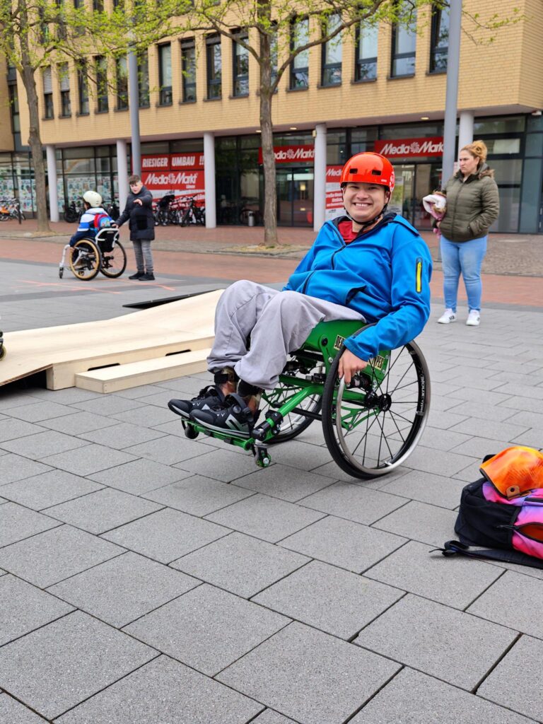 Eine Person mit rotem Helm und blauer Jacke sitzt in einem grünen WCMX-Rollstuhl. Sie balanciert auf den beiden großen Hinterrädern, während die kleinen Vorderräder in der Luft schweben. Die Person lächelt in die Kamera. Im Hintergrund steht eine flache Holzrampe und ein weiteres Kind im Rollstuhl. Die Szene spielt sich auf einem offenen Platz in der Stadt ab. Die Atmosphäre wirkt aktiv und fröhlich.