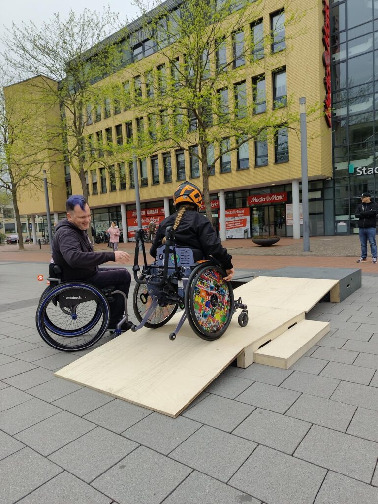 Ein Kind mit orangem Helm fährt in einem Rollstuhl mit bunter Radverkleidung eine breite, flache Holzrampe hinauf. Links neben dem Kind rollt ein Erwachsener im manuellen Rollstuhl mit lila Haaren mit und gibt hilfreiche Tipps. Beide blicken nach vorn zur Rampe. Die Rampe führt auf eine kleine Plattform, die am Rand eines Platzes aufgebaut ist. Im Hintergrund stehen Bäume und ein größeres Gebäude mit gelber Klinkerfassade. Die Stimmung ist konzentriert und ermutigend.