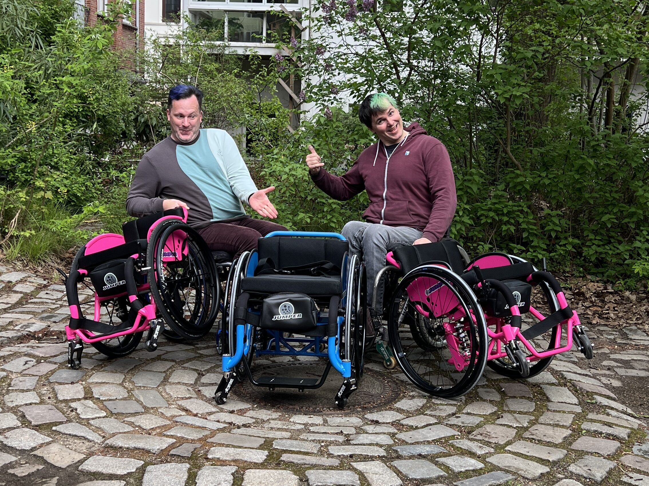 Zwei lächelnde Personen sitzen im Rollstuhl auf einem gepflasterten Platz, umgeben von blühenden Sträuchern. Vor ihnen stehen drei neue WCMX-Rollstühle: zwei in Pink-Schwarz und einer in Blau-Schwarz. Es ist ein freundlicher Frühlingstag und die Stimmung ist gelöst.