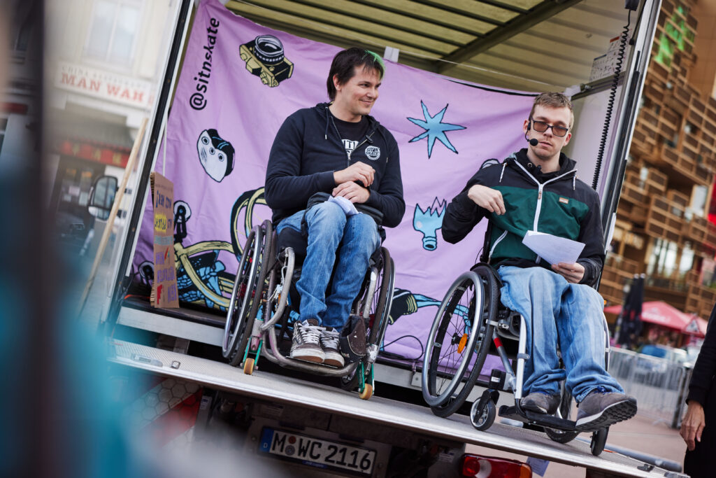Zwei Menschen im Rollstuhl auf einer mobilen Bühne sprechen in Mikrofone.