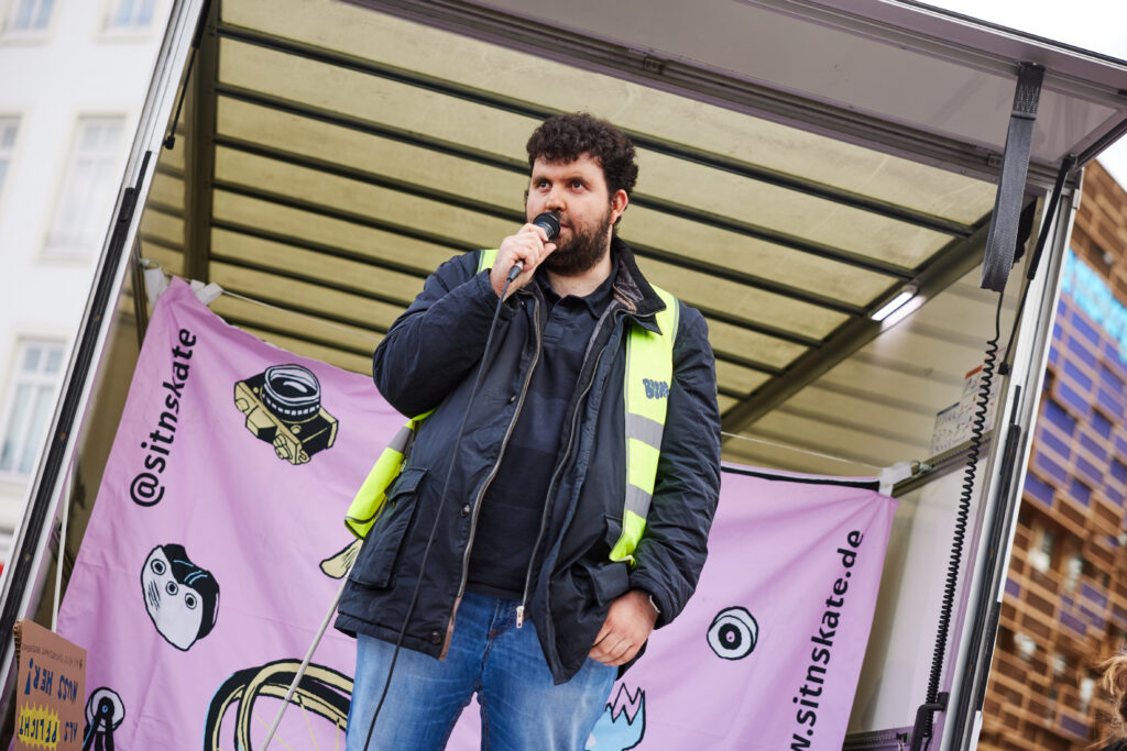 Eine Person spricht energisch in ein Mikrofon, stehend auf der Bühne, vor einem SIT’N’SKATE-Banner.
