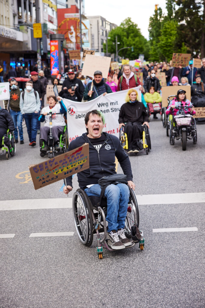 Foto von einem Demozug auf der Reeperbahn, ein Rollstuhlfahrer fährt vorn weg und schreit. Er hält ein Schild in der Hand auf dem steht: Kein Vertrösten mehr, Barrierefreiheit als Pflicht muss her! Dahinter viele Menschen mit und ohne Rollstuhl, mit Schildern und Bannern. 