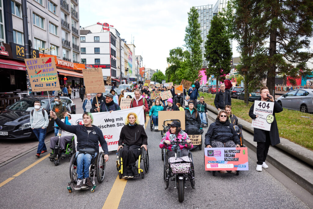 Menschen in Rollstühlen und zu Fuß demonstrieren mit Schildern.