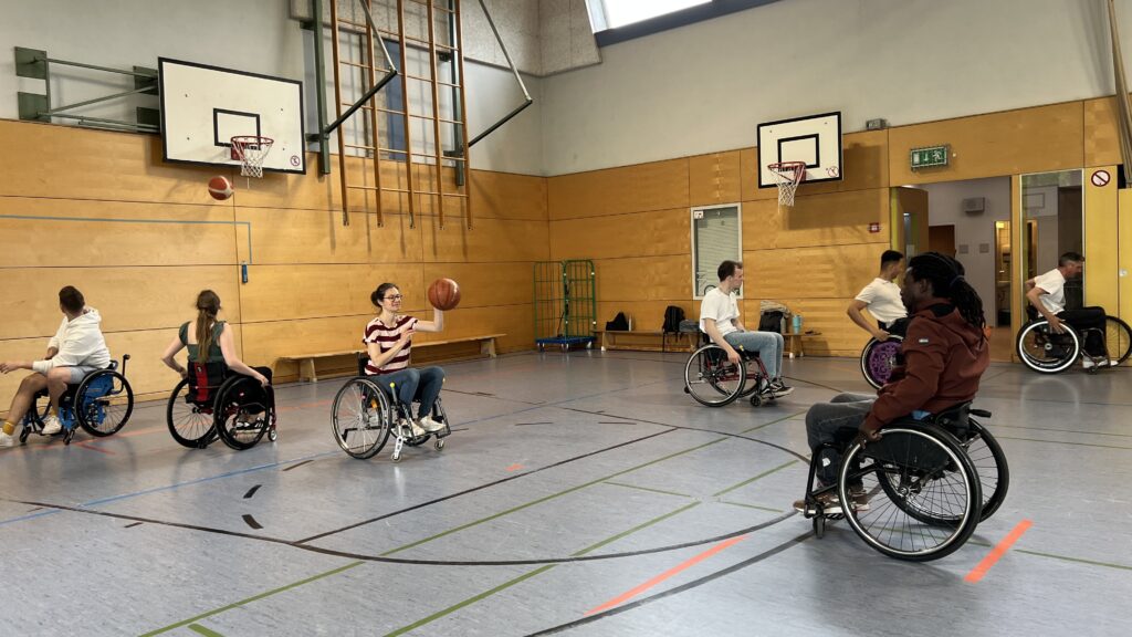 Mehrere Personen im Rollstuhl spielen Rollstuhlbasketball in einer Turnhalle. Eine Frau im Vordergrund fängt gerade den Ball, während andere in Bewegung sind. Es herrscht eine lebendige, sportliche Atmosphäre.