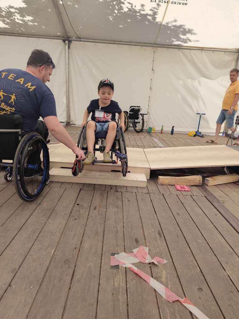 Ein kleiner Junge im Rollstuhl fährt über eine schmale Holzrampe. Ein Erwachsener im Rollstuhl unterstützt ihn dabei. Sie befinden sich in einem weißen Festzelt mit Holzfußboden, im Hintergrund beobachten weitere Personen die Aktion.