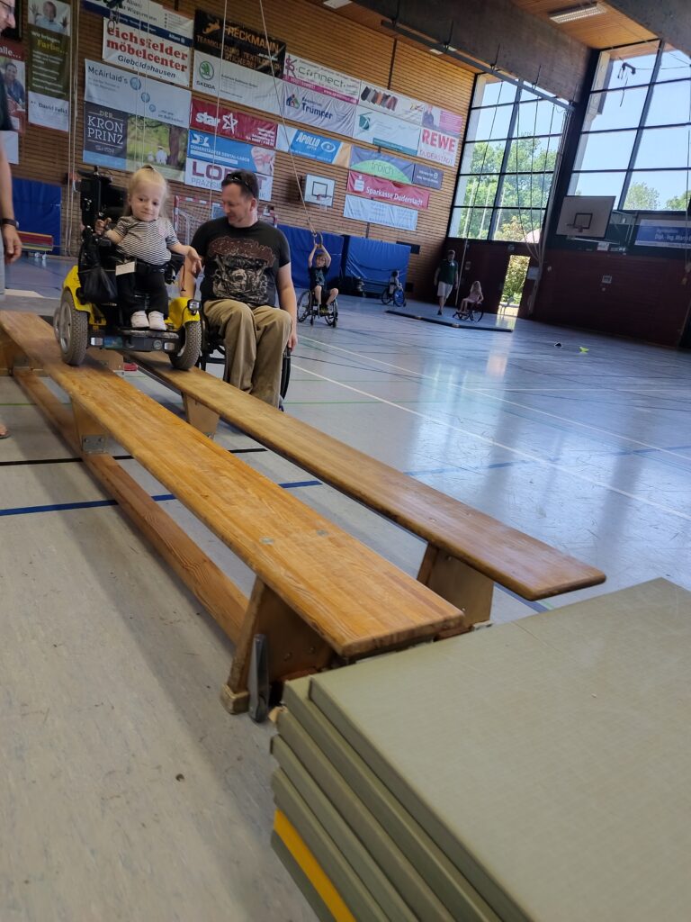 Ein junges Mädchen im E-Rollstuhl fährt vorsichtig über eine Holzbrücke, die aus zwei Turnbänken besteht. Daneben begleitet ein erwachsener Mann im Rollstuhl die Fahrt. Im Hintergrund sind weitere Kinder und Jugendliche in einer großen Sporthalle zu sehen.