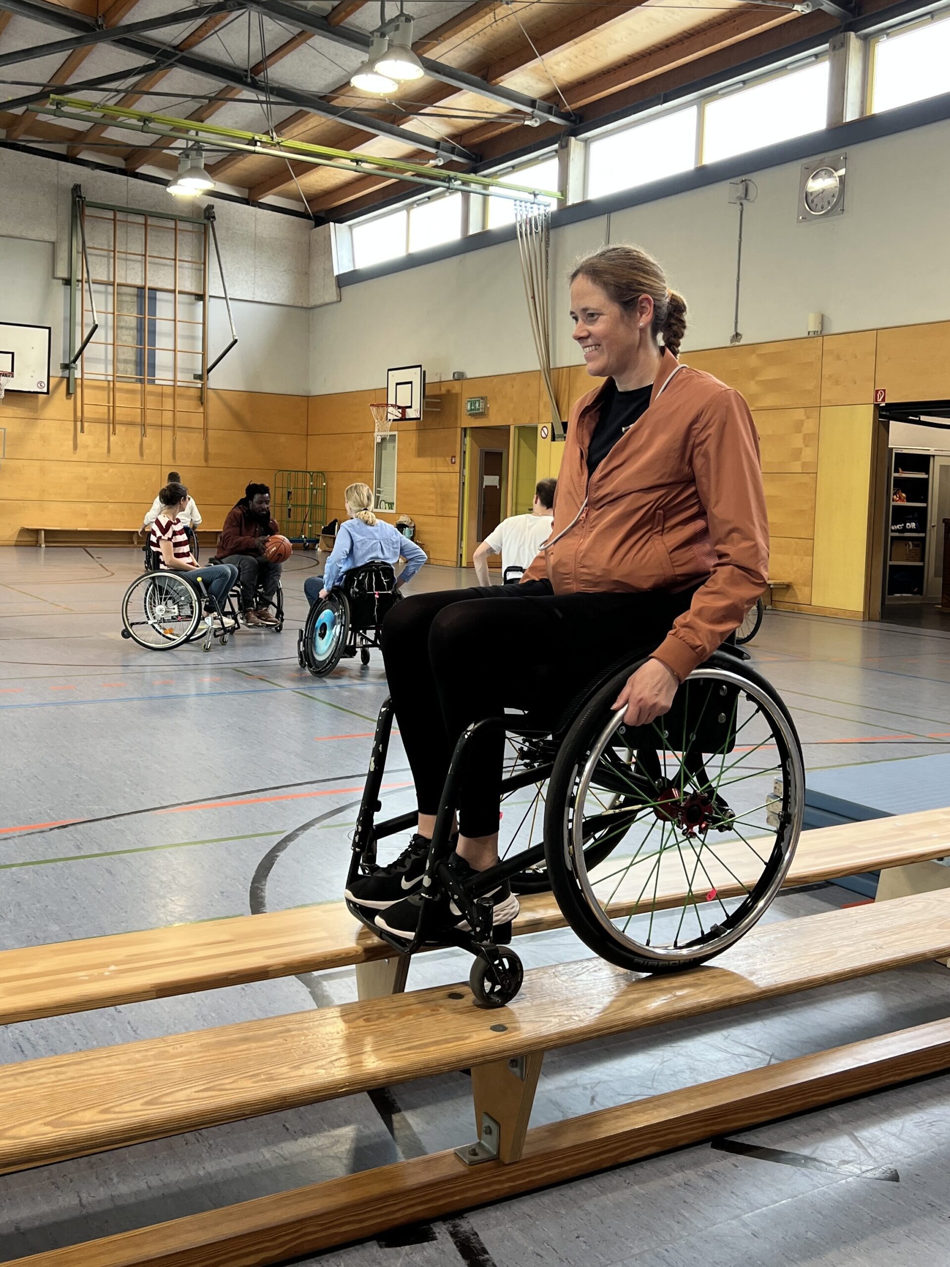 Eine Frau im Rollstuhl balanciert konzentriert über eine Turnbank. Sie trägt eine rostrote Jacke und lacht, während sie mit dem Vorderrad präzise auf dem Holz balanciert. Im Hintergrund üben weitere Teilnehmende in der Sporthalle.