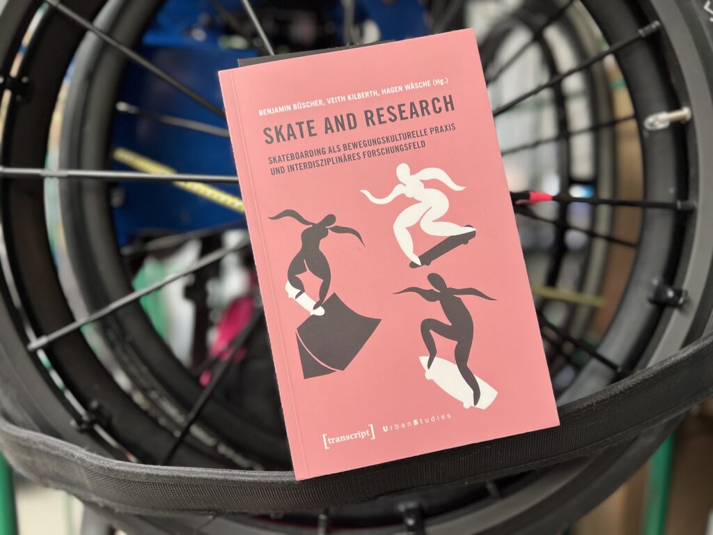 Das Buch Skate and Research mit rotem Cover lehnt auf einem schwarzen Rollstuhlrad. Auf dem Cover sind stilisierte schwarze und weiße Figuren beim Skateboarden abgebildet. Im Hintergrund erkennt man unscharf weitere Teile eines Rollstuhls und bunte Elemente.
