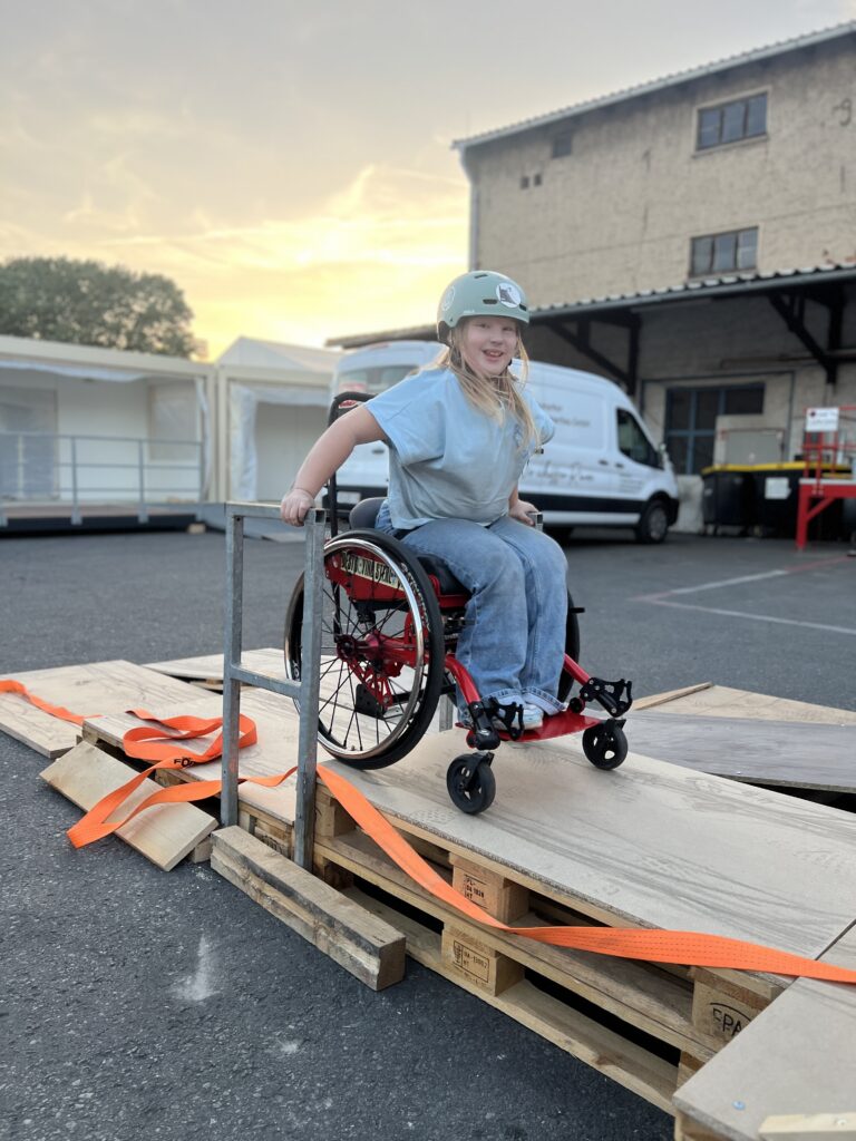 Ein Mädchen mit Helm fährt lächelnd im Rollstuhl über eine improvisierte Rampe aus Holzplatten und Paletten. Der Himmel ist abendrot, im Hintergrund stehen Zelte und Transporter.