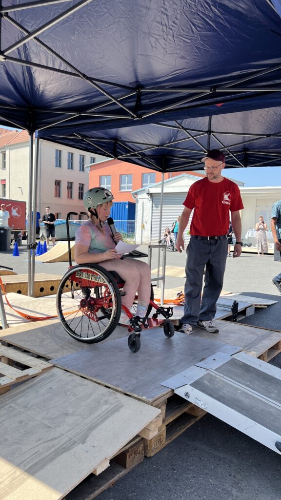 Eine junge Rollstuhlfahrende mit Helm sitzt unter einem blauen Pavillon auf einer Rampe aus Paletten und spricht in ein Mikrofon. Neben ihr steht ein Mann im roten T-Shirt mit Aufdruck “Crew” und beobachtet die Szene aufmerksam.