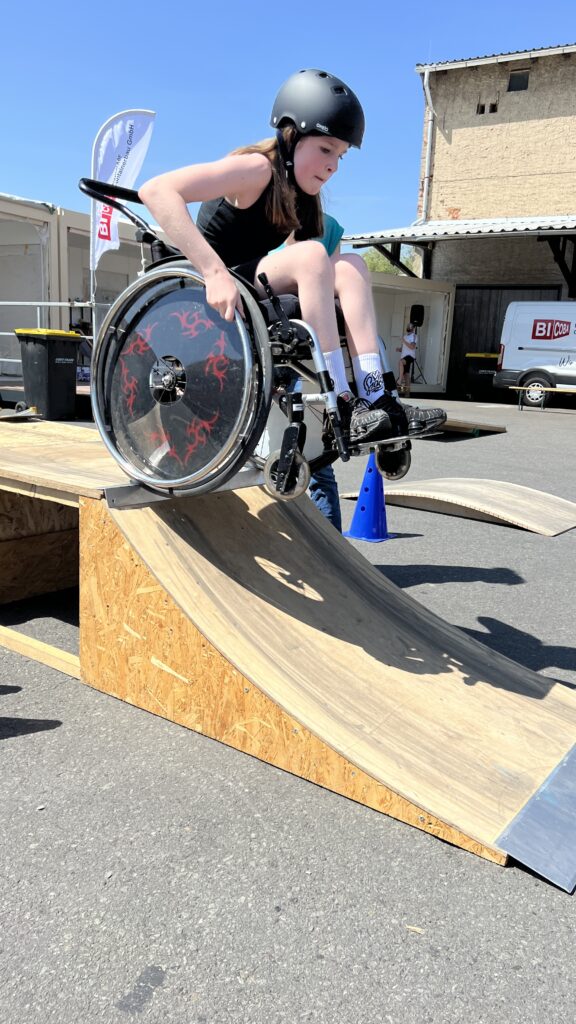 Ein Mädchen im schwarzen Outfit und mit Helm fährt konzentriert mit dem Rollstuhl eine steile Holzrampe im Skatepark hinunter. Der Moment zeigt den Übergang von der Kante zur Schräge – mitten in der Bewegung.