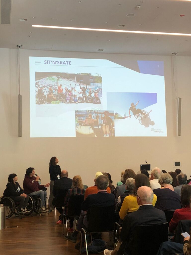 Lisa und David halten gemeinsam einen Vortrag vor einem großen Publikum. David sitzt im Rollstuhl mit Mikrofon, Lisa steht neben ihm. Im Hintergrund eine Präsentation, das Publikum ist aufmerksam und der Saal gut gefüllt.