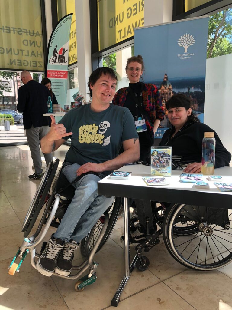 Lisa und David Lebuser stehen mit Nora Gerken von der Bürgerstiftung Hannover an einem Infostand von SIT’N’SKATE. Auf dem Tisch liegen Flyer und Infomaterial, im Hintergrund erkennt man weitere Stände des Stiftungstags. Alle drei lächeln und wirken im Gespräch vertieft.