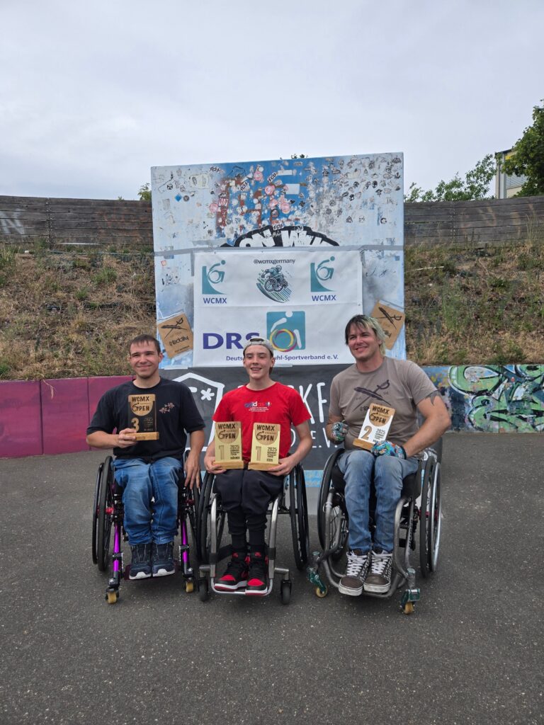 Drei Rollstuhlfahrer posieren lachend mit ihren Pokalen vor einer kleinen Rampe, die mit Bannern der WCMX Germany Open 2025 und des Deutschen Rollstuhl-Sportverbands geschmückt ist. In der Mitte sitzt Max Dorka mit zwei Pokalen für den 1. Platz, rechts neben ihm David Lebuser mit dem Pokal für den 2. Platz, links sitzt der Drittplatzierte mit seinem Pokal. Alle drei strahlen vor einer bunt gestalteten Wand mit Stickern und Graffiti.