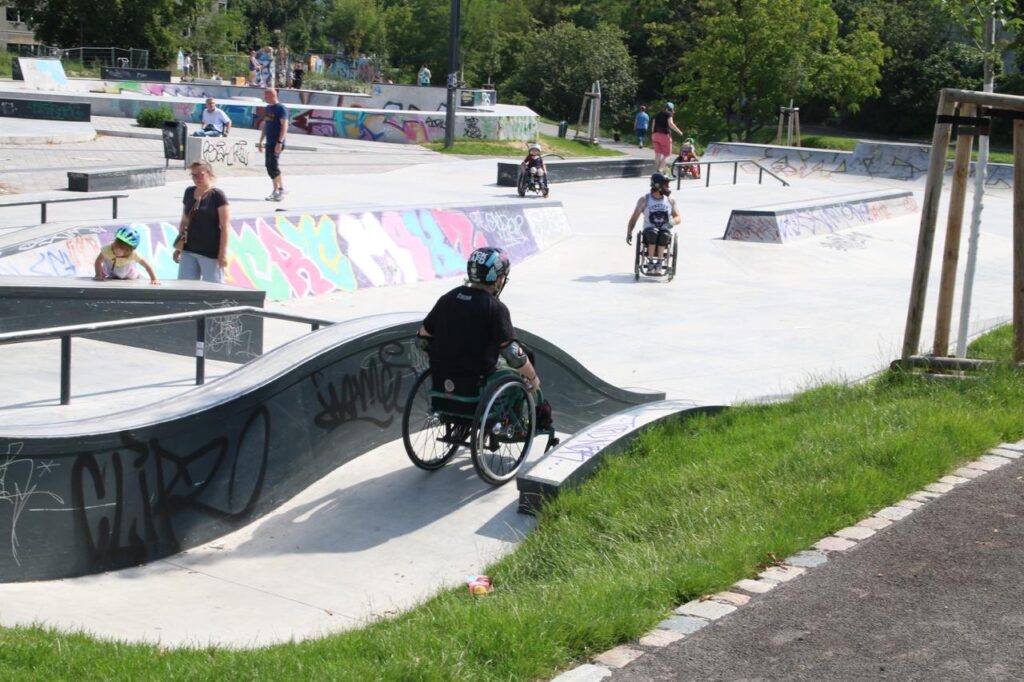 Mehrere Menschen im Rollstuhl bewegen sich durch einen offenen Skatepark. Im Vordergrund rollt eine Person mit Helm eine gebogene Rampe hinunter, im Hintergrund sind bunte Graffitiwände und weitere Teilnehmende zu sehen.