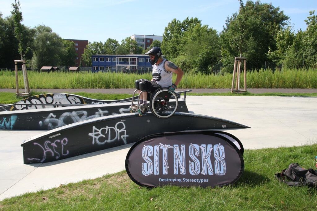 Ein Rollstuhlfahrender mit Helm balanciert auf einem schmalen, geschwungenen Element im Skatepark. Vor ihm steht ein Banner mit dem SIT’N’SKATE-Logo und dem Slogan „Destroying Stereotypes“. Im Hintergrund sieht man Grünflächen und ein Gebäude.