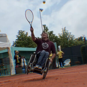 David sitzt in einem Aktivrollstuhl auf einem Sand-Tennisplatz und spielt gerade Rollstuhltennis. Er ist leicht nach hinten gekippt, balanciert auf einem Rad und hat den Tennisschläger erhoben – der Ball fliegt über ihm durch die Luft. David trägt einen weinroten Hoodie mit SIT’N’SKATE-Logo. Im Hintergrund stehen einige Personen am Rand des Platzes, darunter eine Person mit Hut und gelbem Shirt. Der Himmel ist bewölkt.