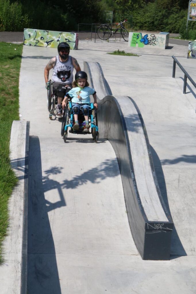 Ein erwachsener Rollstuhlfahrer mit Helm begleitet ein Kind im Rollstuhl eine breite Rampe im Skatepark hinunter. Beide tragen Helme und wirken konzentriert. Im Hintergrund sind weitere Skate-Elemente und Graffiti zu sehen.