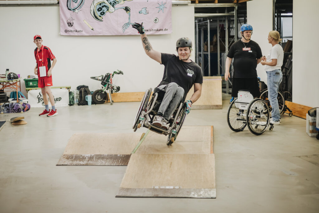 Eine erwachsene Person im Rollstuhl fährt konzentriert auf einem Rad. Ein junger Rollstuhlfahrender fährt mit Schwung über eine Rampe und hebt dabei eine Hand in die Luft. Die Bewegung wirkt dynamisch und sportlich. Weitere Menschen schauen zu oder sprechen im Hintergrund.