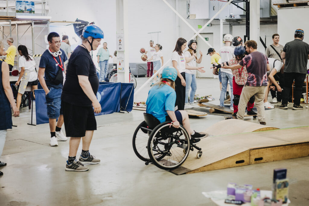 Eine Person sitzt in einem Skate-Rollstuhl auf einer Rampe. Im Hintergrund stehen Menschen und beobachten das Geschehen, fahren Skateboard mit Unterstützung oder spielen Basketball. Die Atmosphäre wirkt sportlich und offen.
