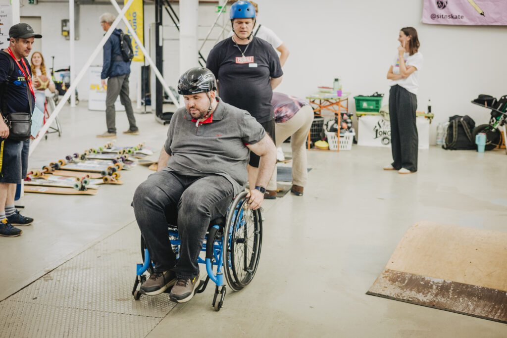 Eine erwachsene Person im Rollstuhl fährt konzentriert mit dem Rollstuhl. Dahinter ein Mann mit Helm. Es stehen weitere Menschen mit Helmen, Skateboards und Rollstühlen im Hintergrund.