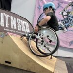 Ein Kind mit Helm fährt in einem Rollstuhl eine kleine Holzrampe hoch. Im Hintergrund sind ein großes lilafarbenes Banner mit einem Rollstuhlskater-Motiv und das SIT’N’SKATE Logo auf einer Beachflag zu sehen. Die Szene spielt in einer Messehalle.